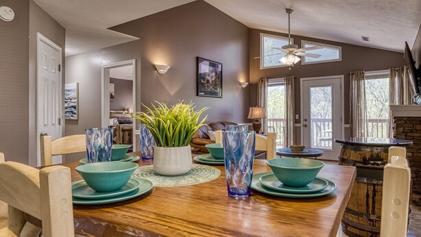 Un coin repas avec une table en bois dressée pour quatre personnes, entourée de chaises. On y trouve deux appliques murales, un ventilateur de plafond et une cheminée avec un manteau en pierre.