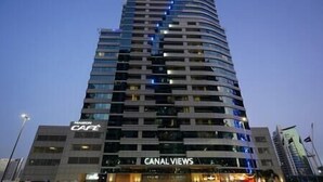 A multi-story hotel with a curved facade, featuring a 'JAMAC' sign at the top, a 'CANAL VIEWS' sign at the bottom, and a 'CAFE' sign on the left. - Splendid1BRapartmentinCanalViews (Dubai)