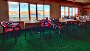 A dining area with tables and chairs, large windows with a view of mountains and a lake, and a patterned ceiling. - World Attic Camp (Leh)