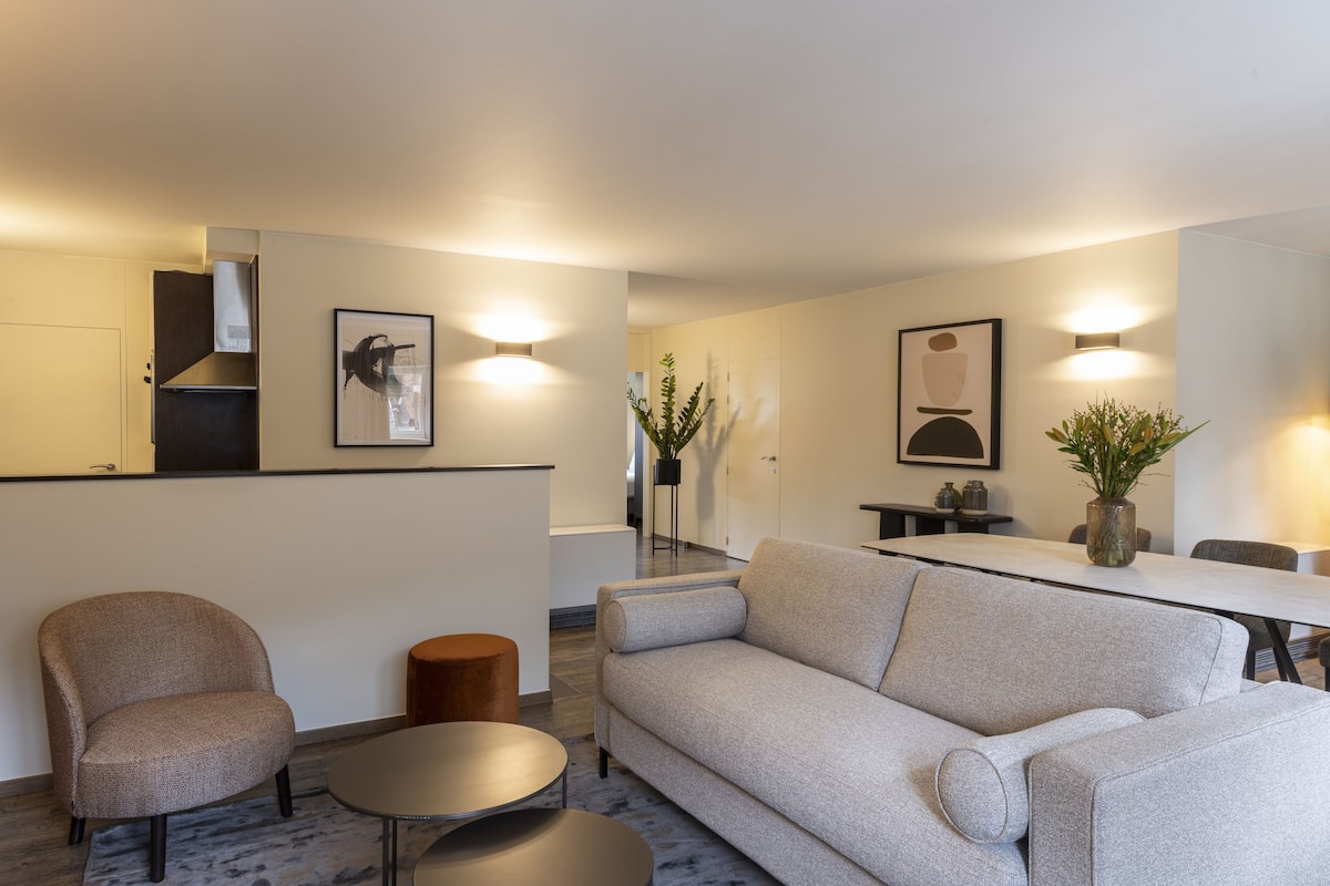 Sala de estar moderna com sofá, poltrona e mesa de centro. Há uma área de jantar com mesa e cadeiras. O ambiente tem uma paleta de cores neutras e apresenta obras de arte e plantas.