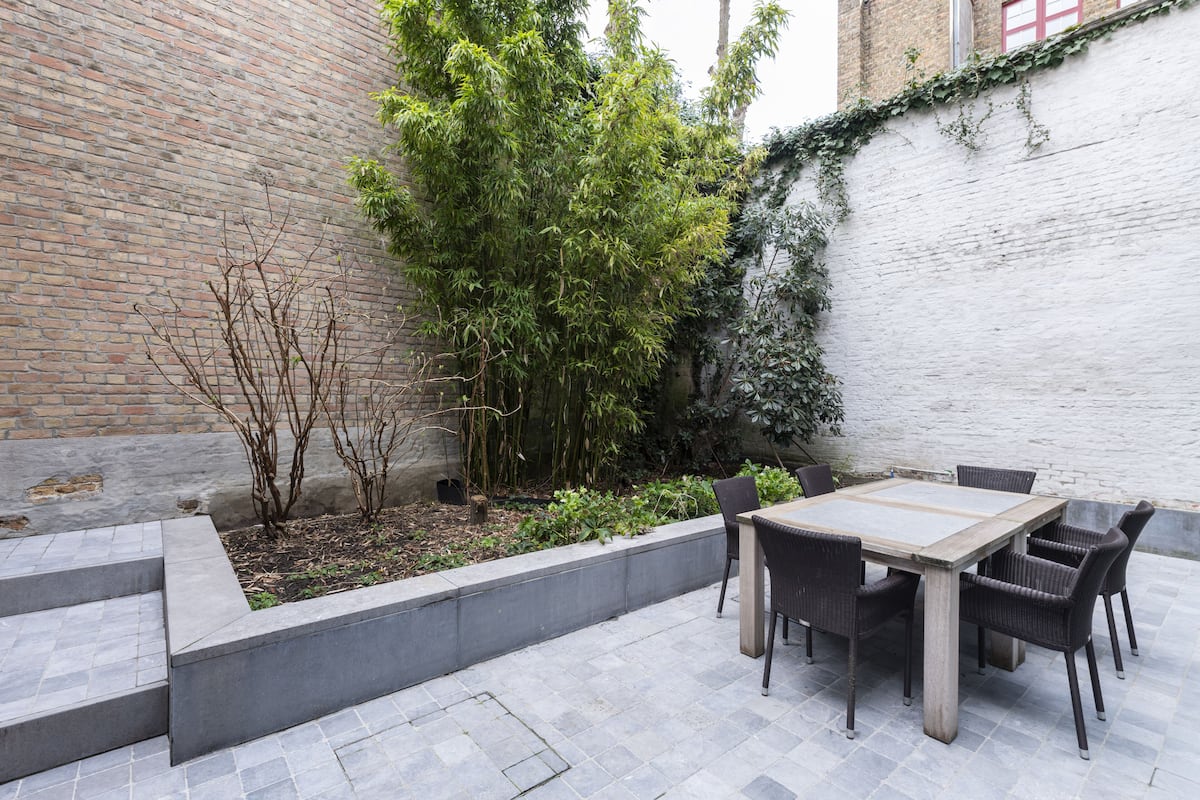 Pátio com mesa e cadeiras, cercado por um muro de tijolos e vegetação.