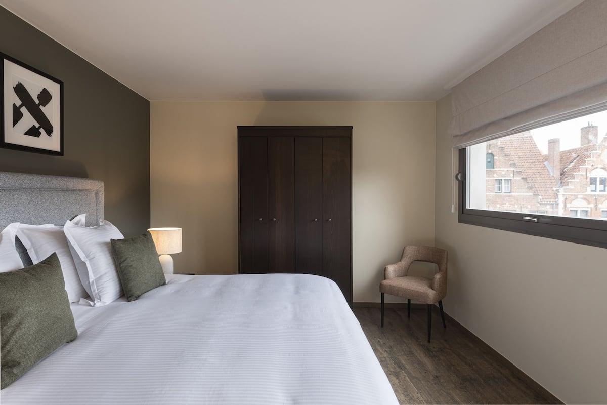 Quarto de hotel com cama, cadeira, guarda-roupa, janela e um quadro fixado na parede.