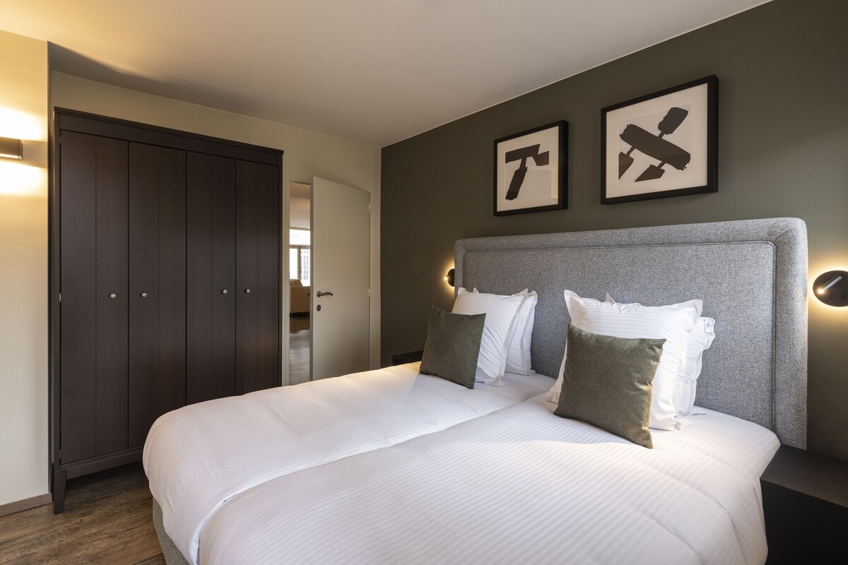 A neatly made bed with a grey headboard, a dark wooden wardrobe, and two framed pictures on the wall.