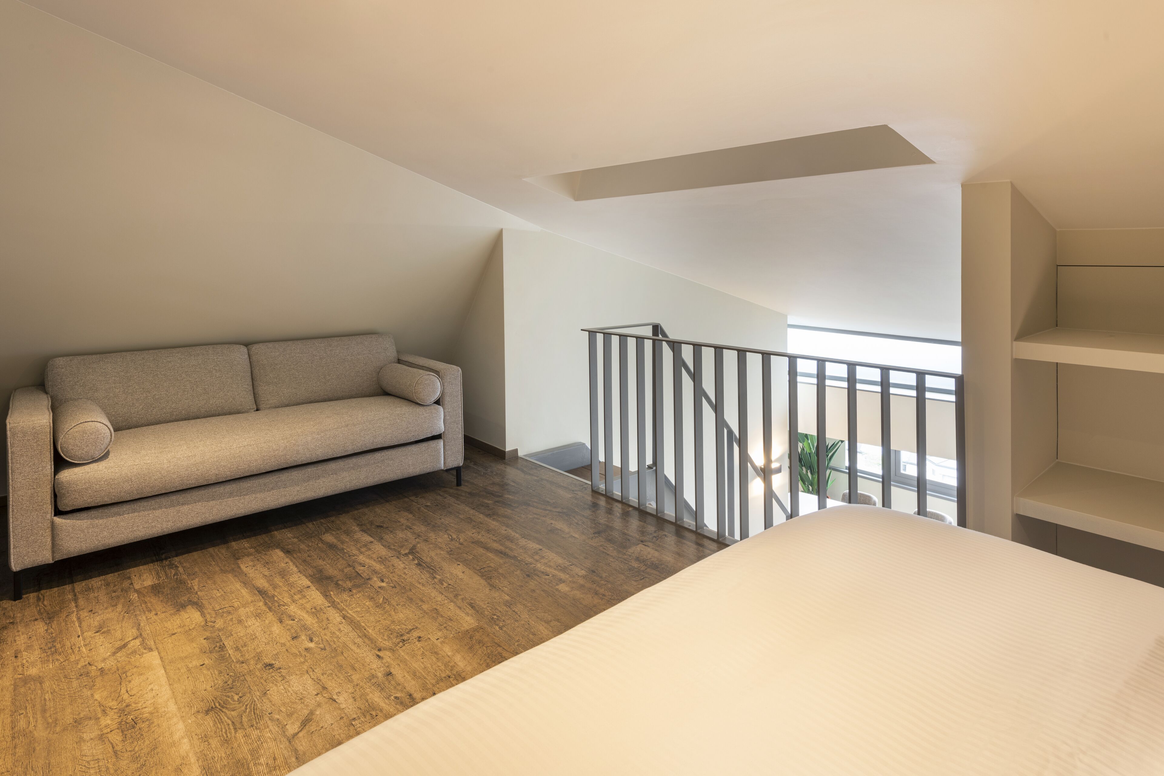 A modern loft bedroom with a wooden floor, a beige sofa, a white bed, and a balcony with a railing.
