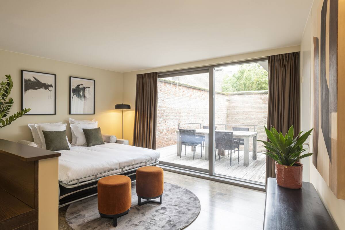 Quarto de hotel moderno com cama, escrivaninha, cadeira, planta em vaso e vista para o pátio externo.
