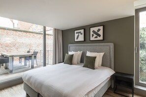 A bedroom with a large bed, a headboard, a bedside table, and a window overlooking a brick wall. - Dukes’ Apartments Grand Place – by Dukes’ Hotel Collection (Bruges)
