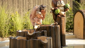 A woman and a child interacting with a wooden squirrel sculpture holding acorns, set against a backdrop of tall grass and a wooden fence.