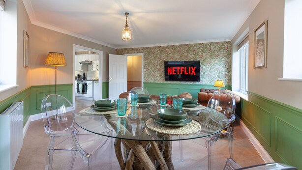 Un área de comedor con una mesa de vidrio, sillas transparentes y un sofá verde. Un televisor muestra "NETFLIX" montado en la pared.