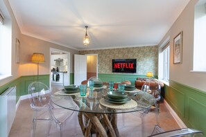 Un coin repas avec une table en verre, des chaises transparentes et un canapé vert. Un téléviseur affichant le logo « NETFLIX » est fixé au mur.