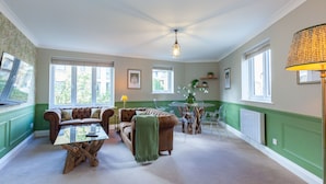 Un salón amplio con una decoración en tonos verde y blanco, que incluye un sofá de cuero marrón, una mesa de centro de vidrio y otra de madera. Hay varias ventanas que dejan entrar luz natural, y se ve una lámpara en el lado derecho.