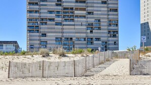 A multi-story building with balconies, a sandy beach in the foreground, and a clear blue sky.