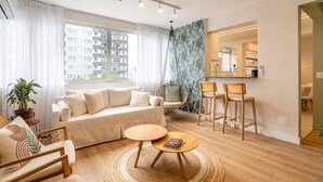 Ein modernes Wohnzimmer mit einer Couch, Sesseln, einem Couchtisch und einem Hängekorbsessel. Es gibt eine Küchenzeile mit einer Arbeitsfläche und Stühlen.