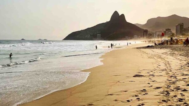 Ein Strand mit einem markanten Berg im Hintergrund, sandigem Ufer und einigen Personen, die am Wasser entlanggehen.