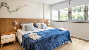 Ein modernes Schlafzimmer mit einem Holz-Kopfteil, einem großen Fenster mit Blick auf Grünflächen, einer blauen Tagesdecke und einem Nachttisch mit Lampe.