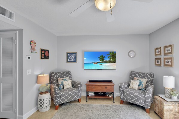 A living room with two patterned armchairs, a flat-screen TV, a wooden coffee table, and a decorative wall clock. - Less than 1/2 mile to beach.  Community pool. (St. Simons Island)