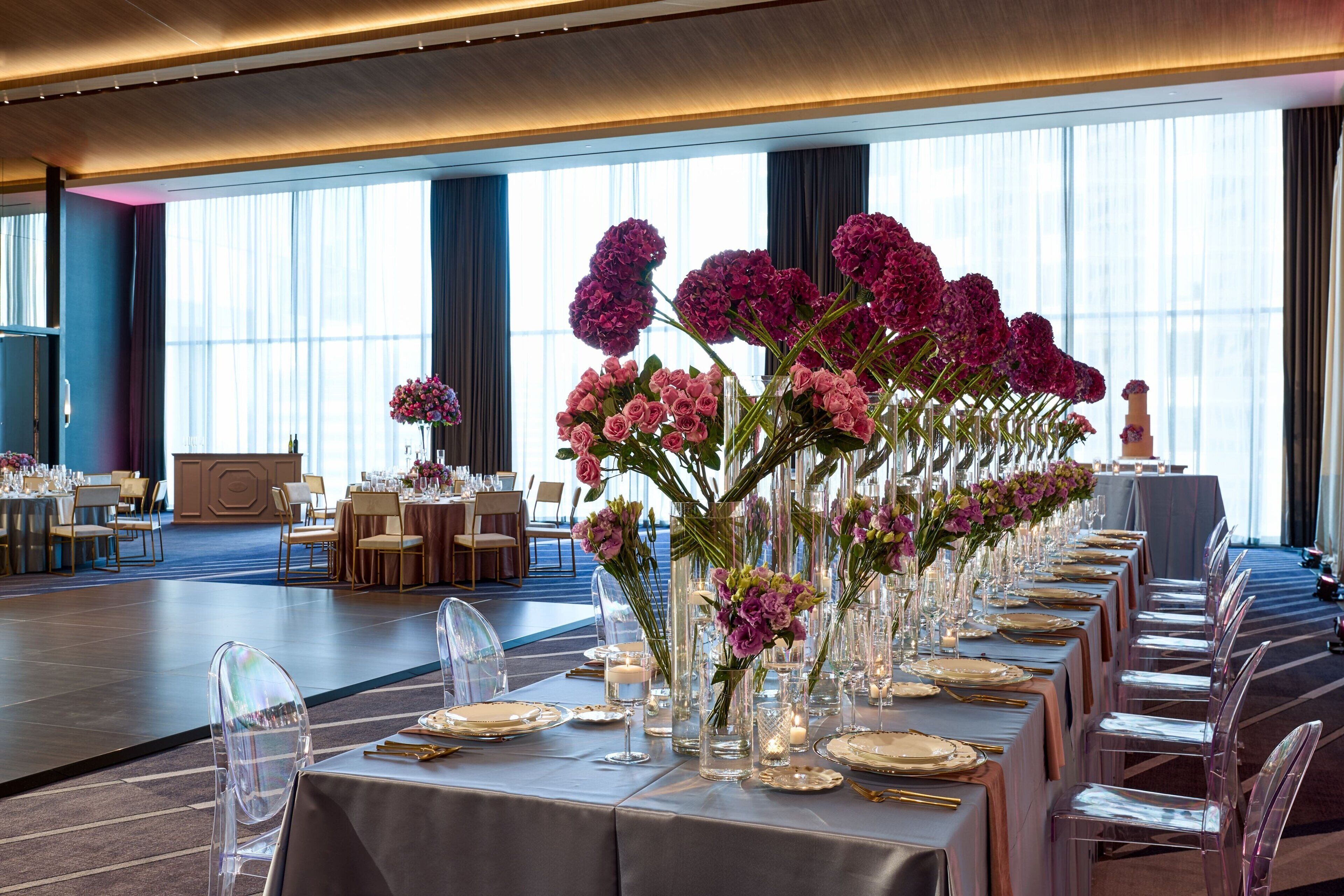 Un salón de banquetes con largas mesas dispuestas para un evento formal, sillas transparentes y centros de mesa florales.