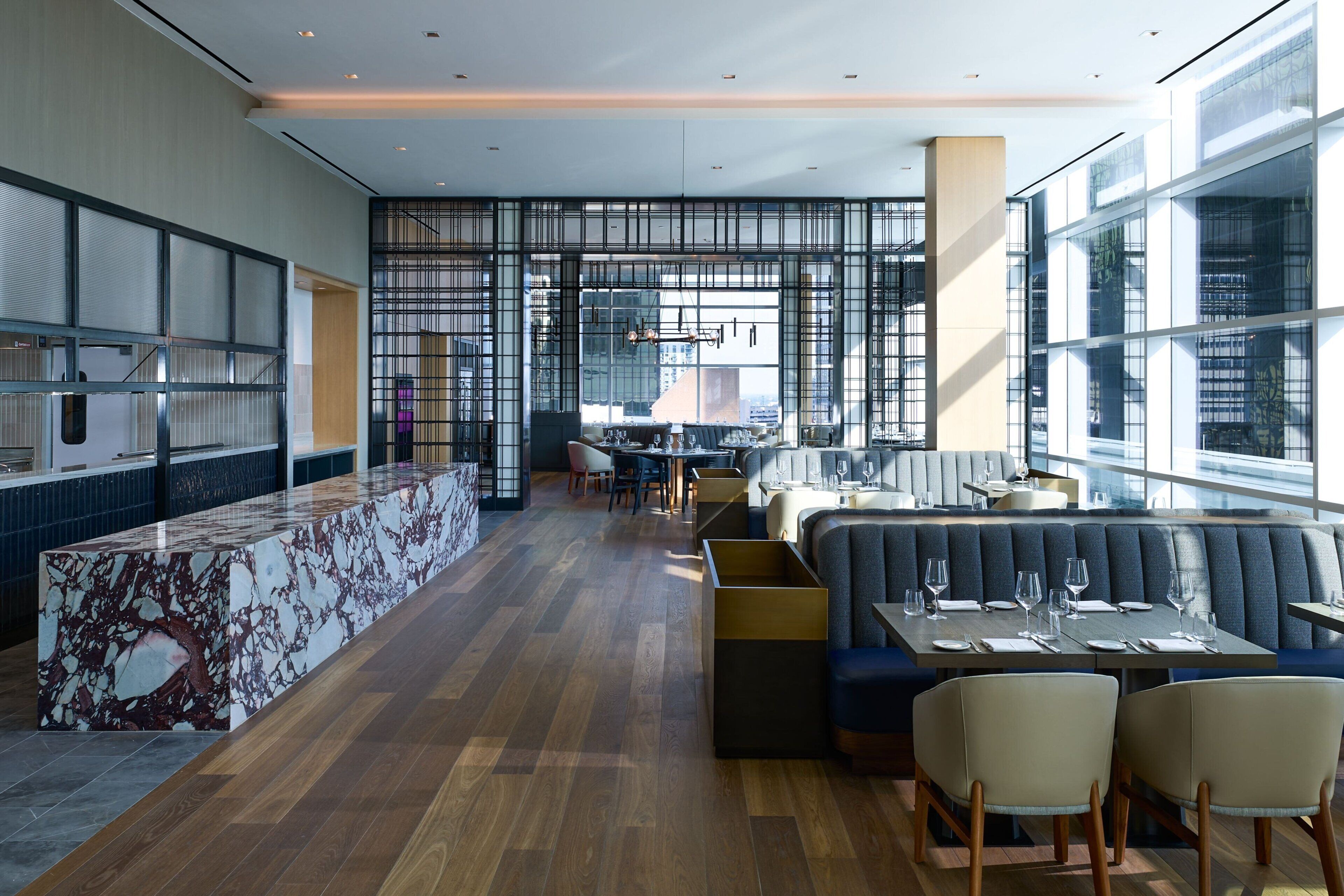A modern restaurant with a marble bar, seating area with sofas and chairs, and large windows offering views of the city.
