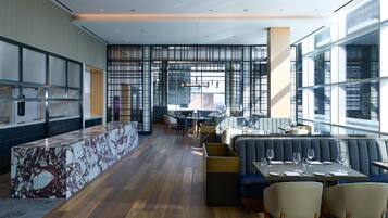 A modern restaurant with a marble bar, seating area with sofas and chairs, and large windows offering views of the city.