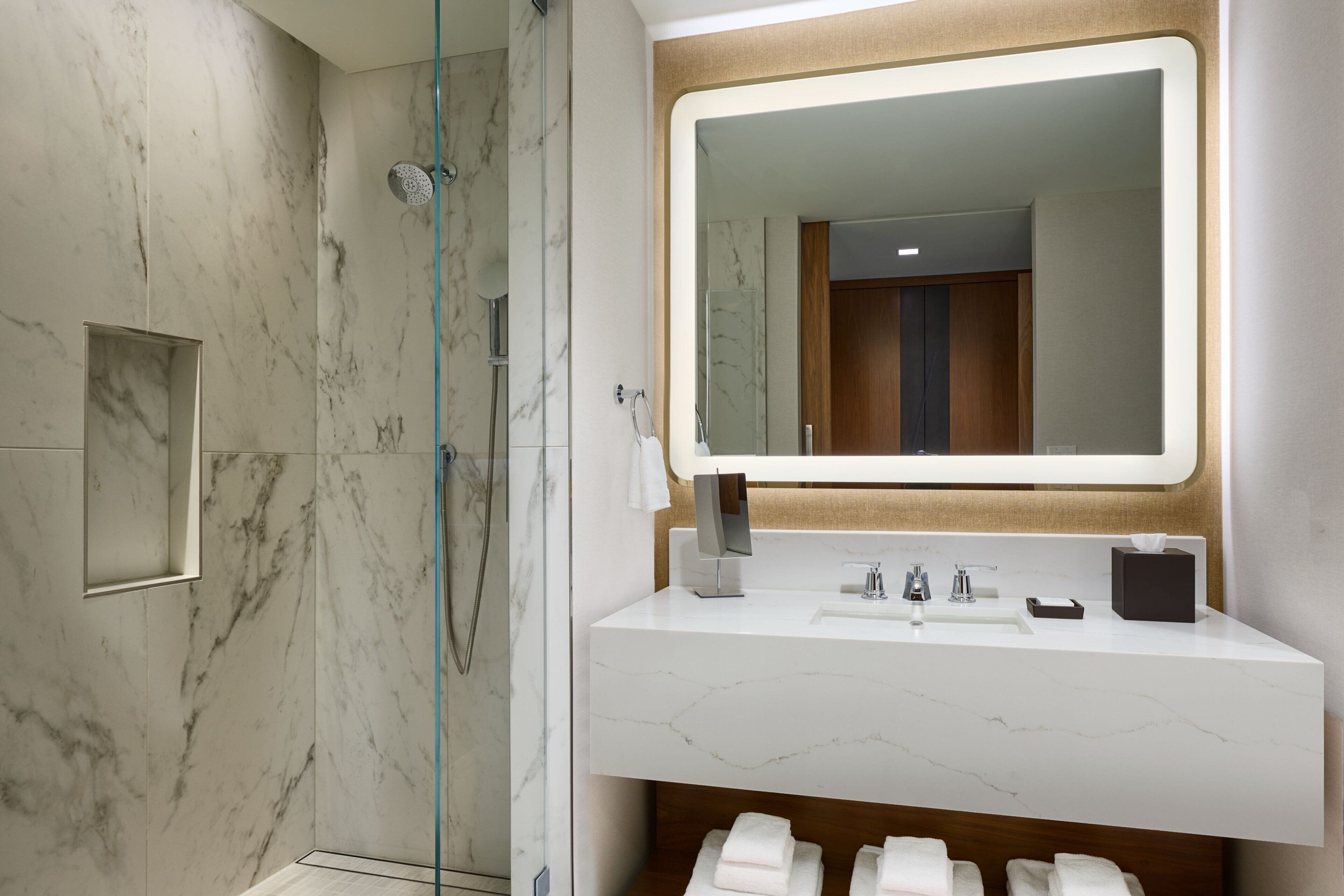 Une salle de bain moderne avec une douche à parois de verre, un mur recouvert de carreaux de marbre, un meuble-lavabo blanc avec un miroir et un meuble en bois en dessous.