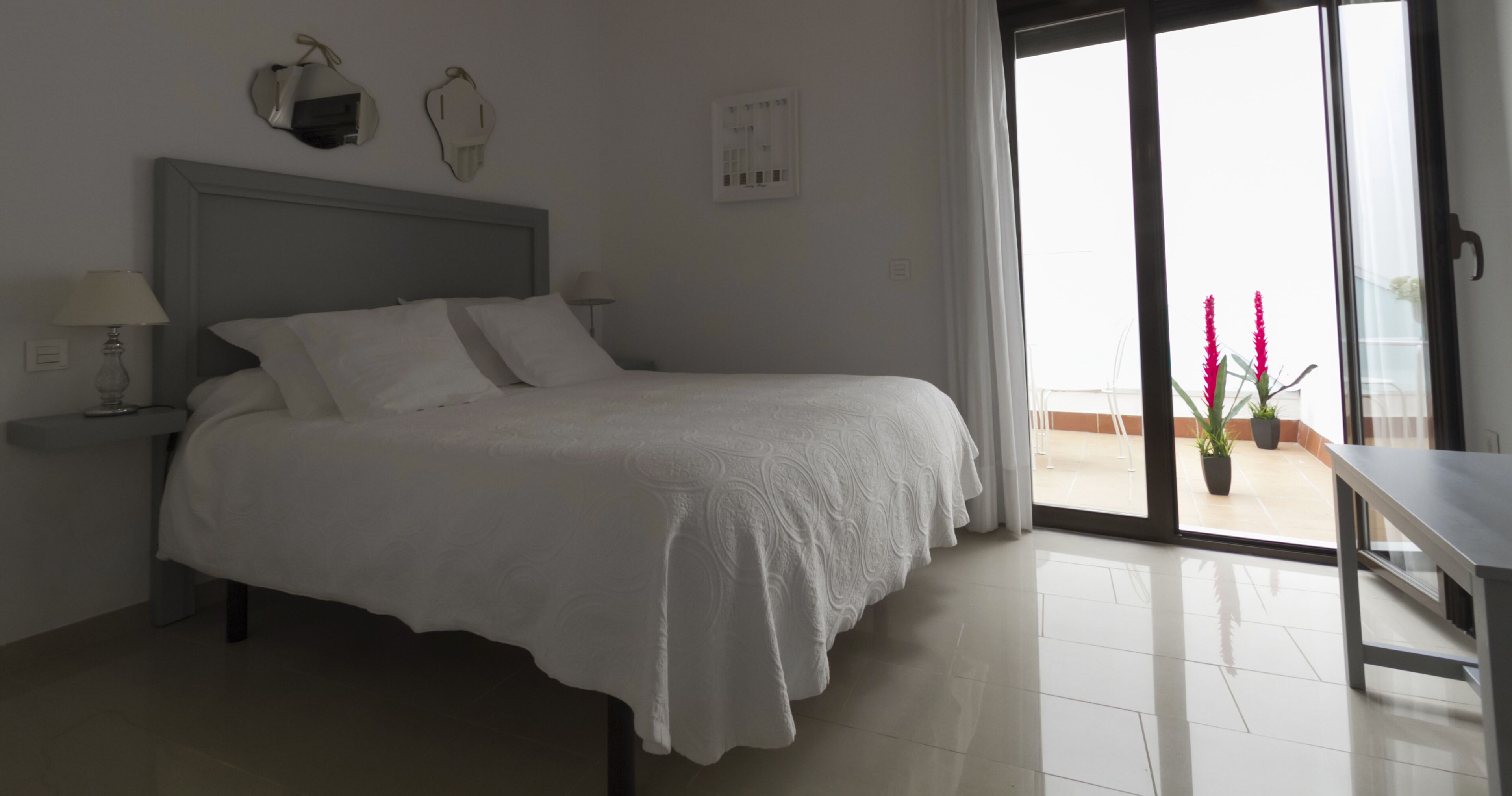 A hotel room with a bed, bedside tables, a window with a view of the outside, and a balcony with potted plants.