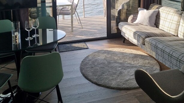Ein modernes Interieur mit einer Glastür, die auf einen Balkon mit Blick auf das Wasser führt. Es gibt eine Sitzecke mit einer Couch und Sesseln, einen kleinen Tisch mit Gläsern sowie einen Fernseher auf einem Lowboard.