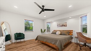 Ein Schlafzimmer mit einem Bett, einem Deckenventilator, einem Sessel, einer Kommode und zwei Fenstern mit Jalousien.