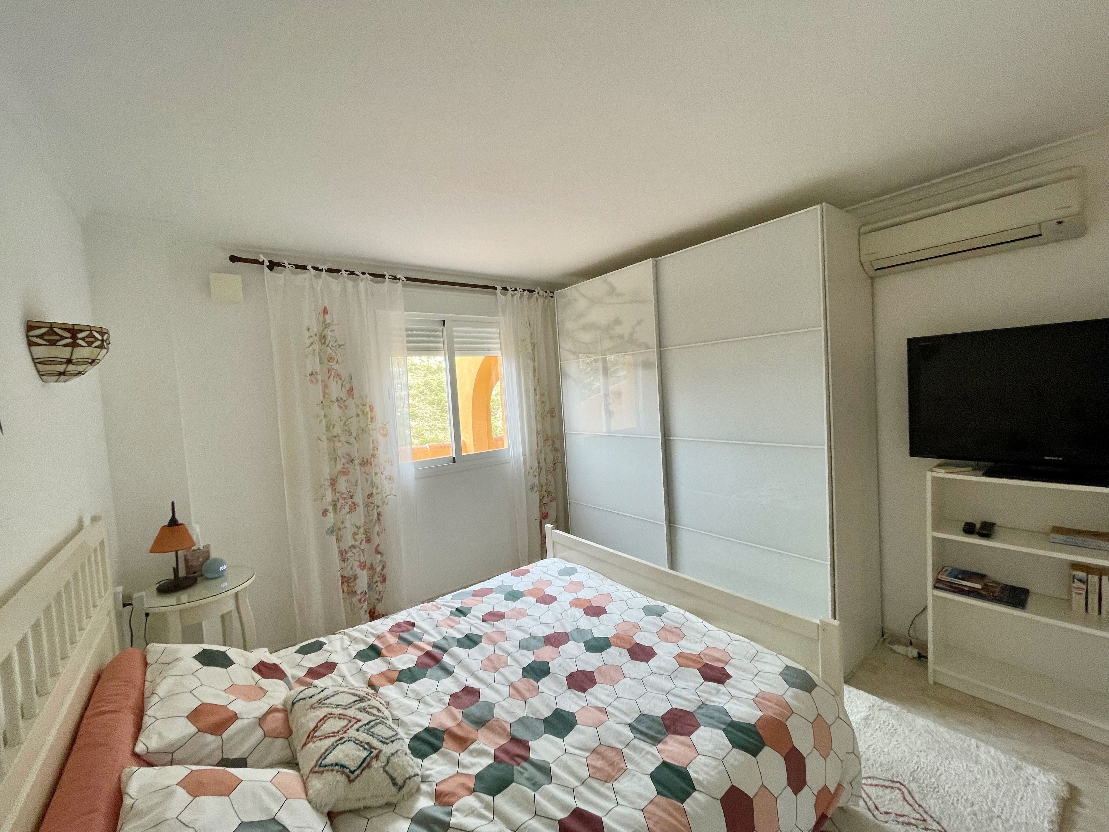 Quarto com cama, televisão em móvel, janela com cortinas e pequena mesa com abajur.