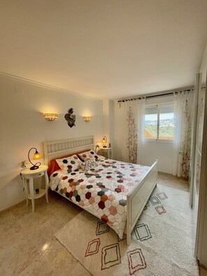 Un dormitorio con cama, dos mesitas de noche, una lámpara de pared y una ventana con cortinas.