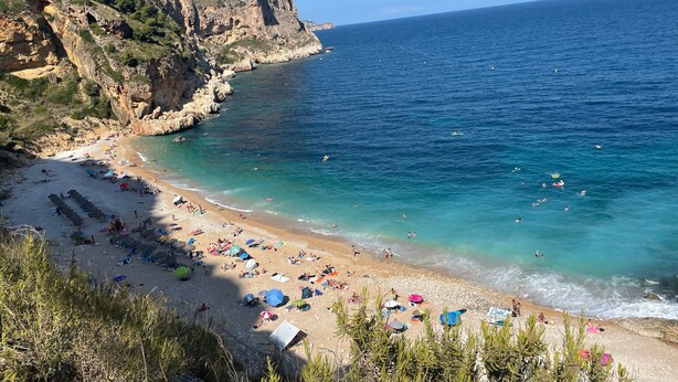 Una playa costera con acantilados rocosos, mar azul cristalino y sombrillas.