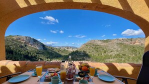 Una mesa de comedor dispuesta con platos, vasos y diversas opciones de comida, con vistas a un paisaje montañoso.