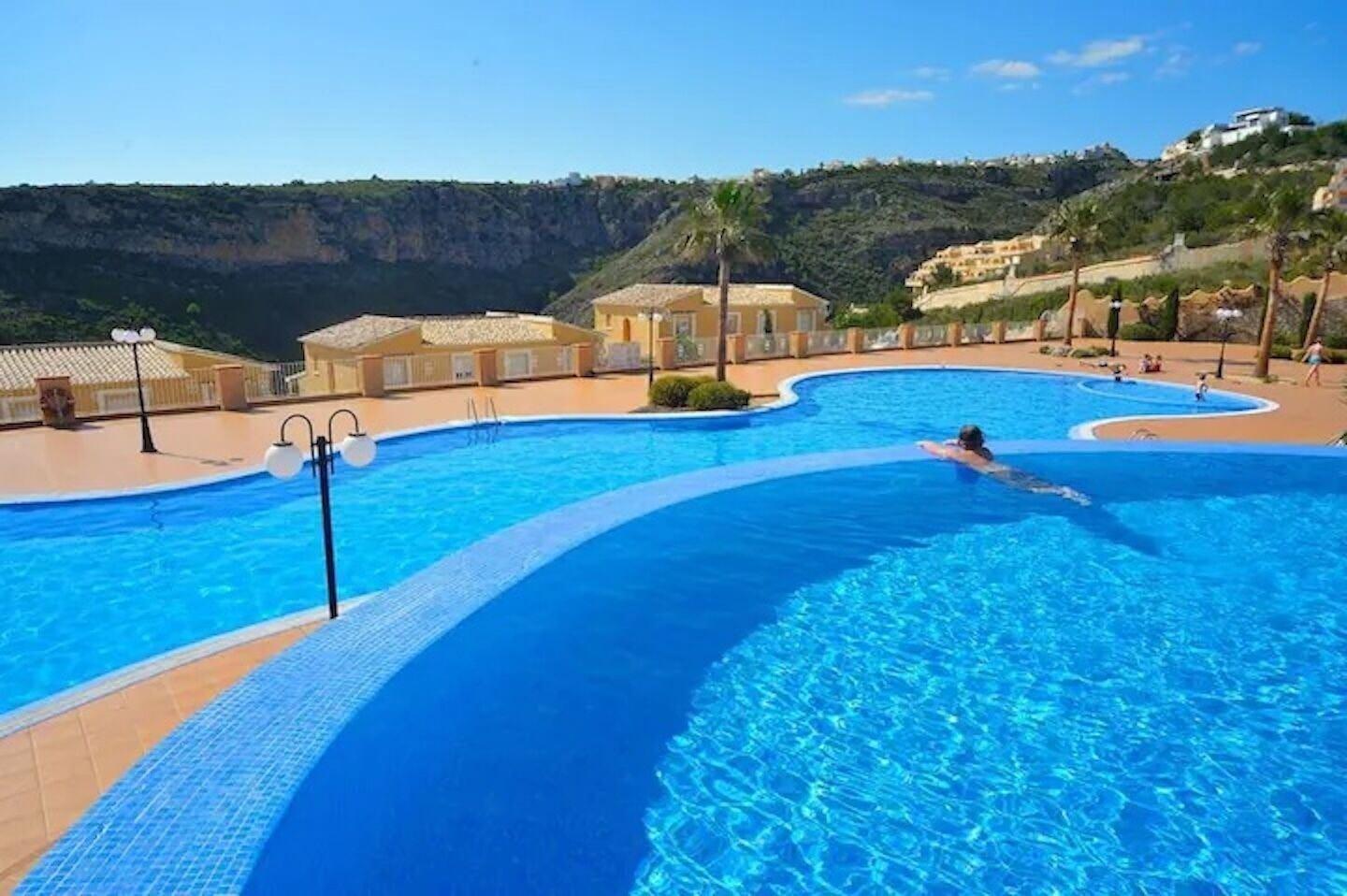 A large outdoor swimming pool with a curved design, surrounded by buildings and a hilly landscape.