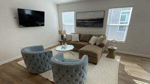 Ein Wohnzimmer mit einem Ecksofa, zwei gesteppten Poufs, einem Couchtisch, einem flachbildfernseher an der Wand und zwei Fenstern mit Jalousien.