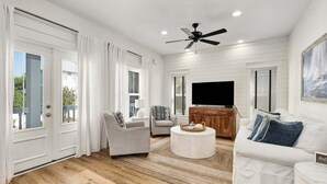 A spacious living room with a ceiling fan, a flat-screen TV, and a large window with white curtains.