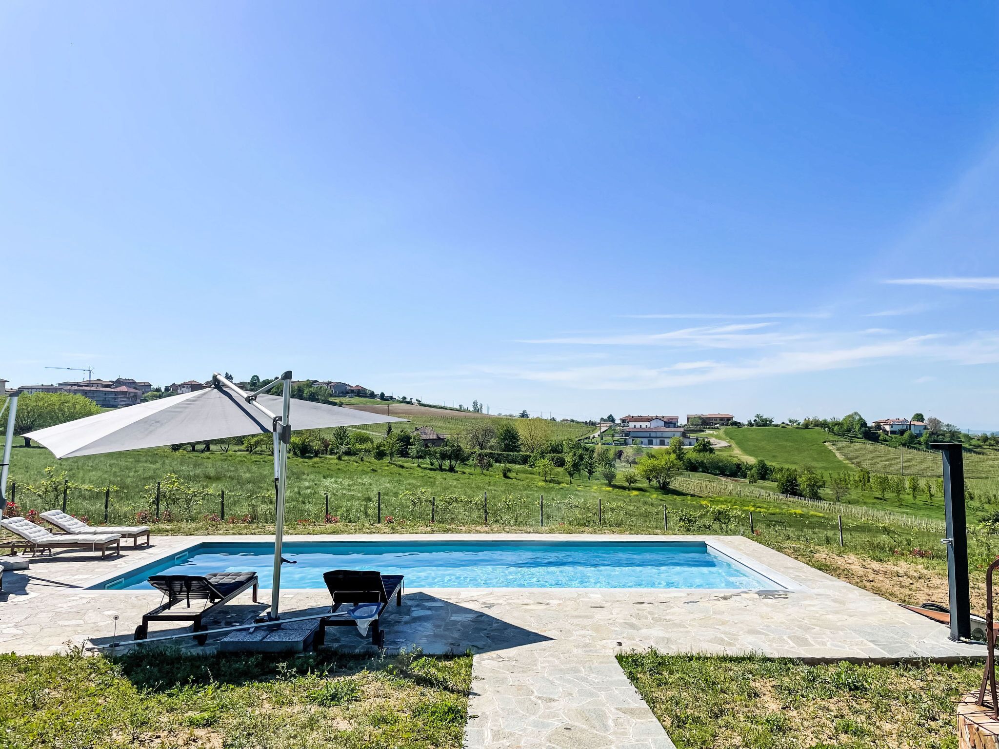 A swimming pool with lounge chairs and a pergola, surrounded by a grassy landscape and houses in the background.