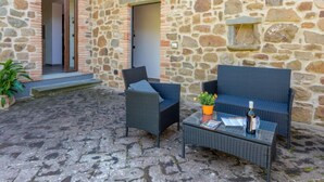 En bygning i sten med en terrasse, møbleret med fletmøbler og et glasbord.