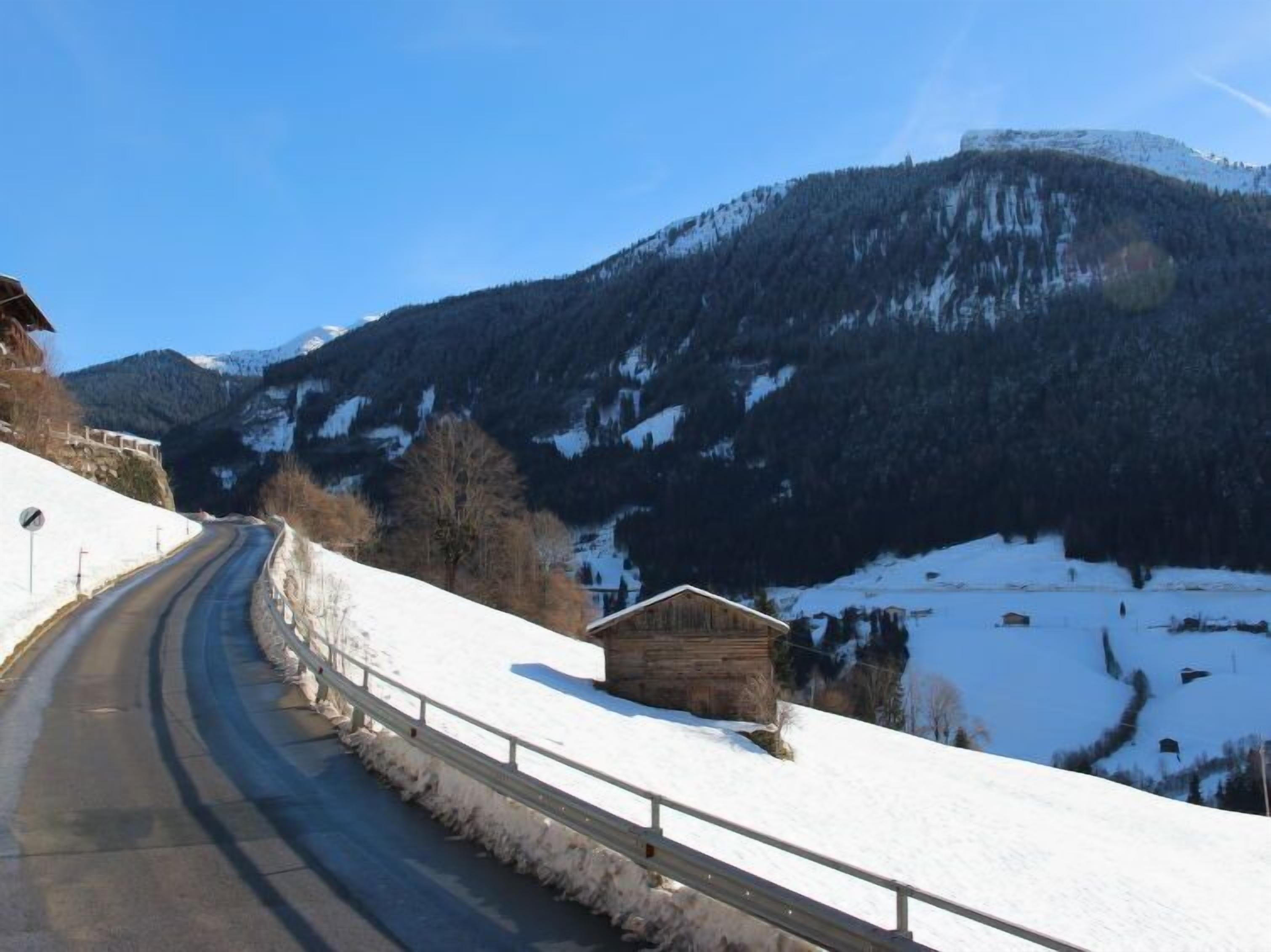 A snow-covered road curving through a mountainous area with a wooden cabin on the right.