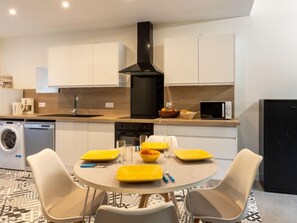 A modern kitchen with white cabinets, a black range hood, a microwave, and a dishwasher. There is a dining table with yellow placemats and a bowl of fruit.