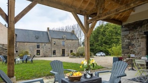 Un espace extérieur aménagé en terrasse, avec des meubles en osier, une table sur laquelle se trouve un panier de fruits, et un bâtiment en pierre en arrière-plan.