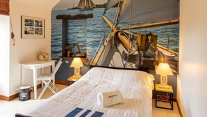 Une chambre avec un lit, deux tables de chevet avec des lampes, un petit bureau et une chaise. Un grand tableau représentant un voilier est accroché au mur.
