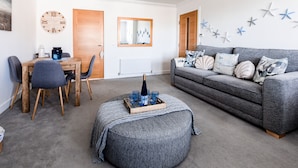 A cozy living room with a grey sofa, a wooden dining table with chairs, a wooden coffee table with a bottle and glasses, and a grey ottoman. - Jules By The Sea Anstruther (Anstruther)