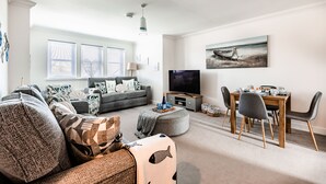 A modern living room with a grey sofa, a dining table with chairs, a flat-screen TV, and a painting of a boat on the wall. - Jules By The Sea Anstruther (Anstruther)