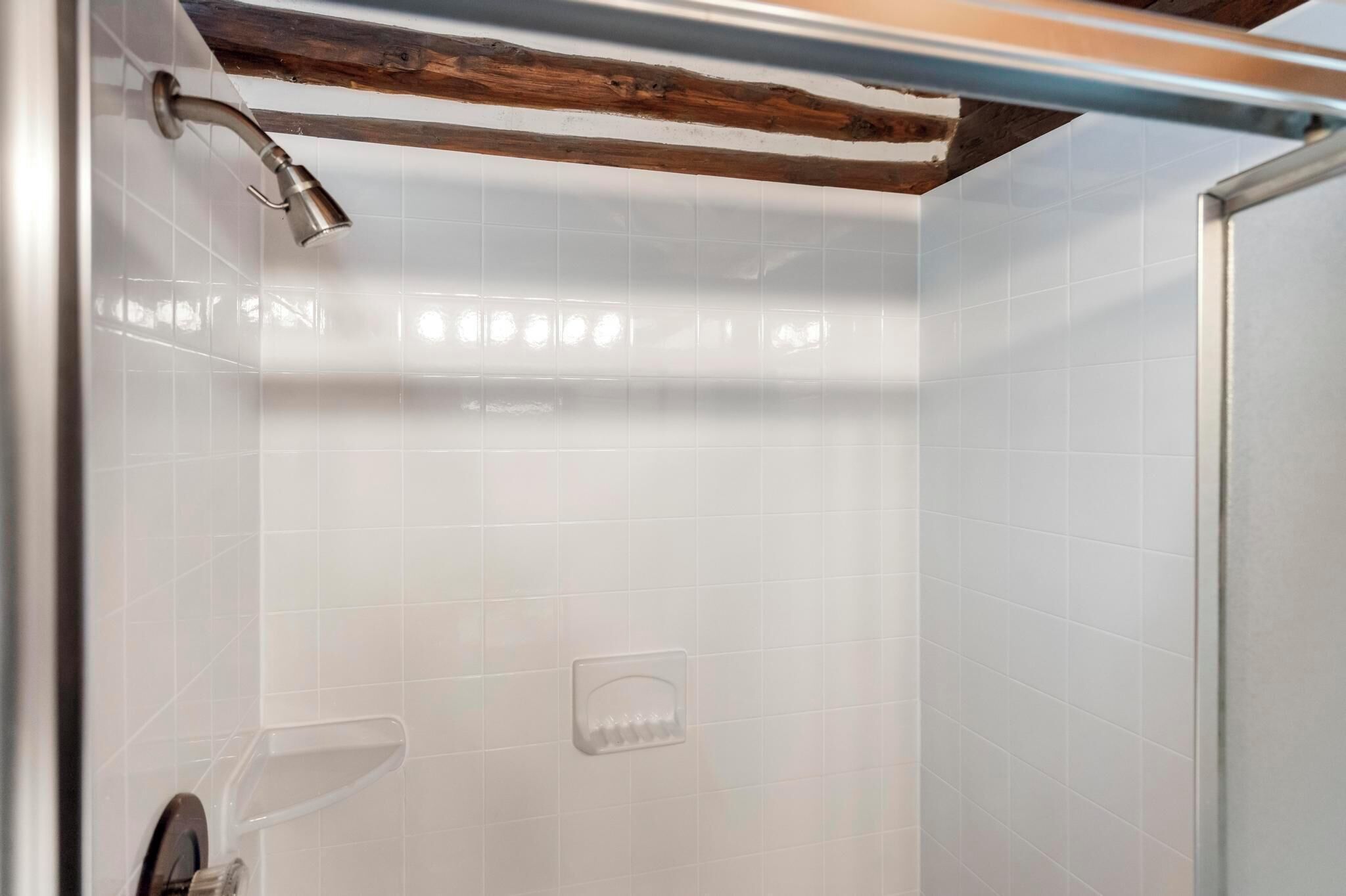 A shower with white tiles, a wooden beam ceiling, and a clear shower caddy.