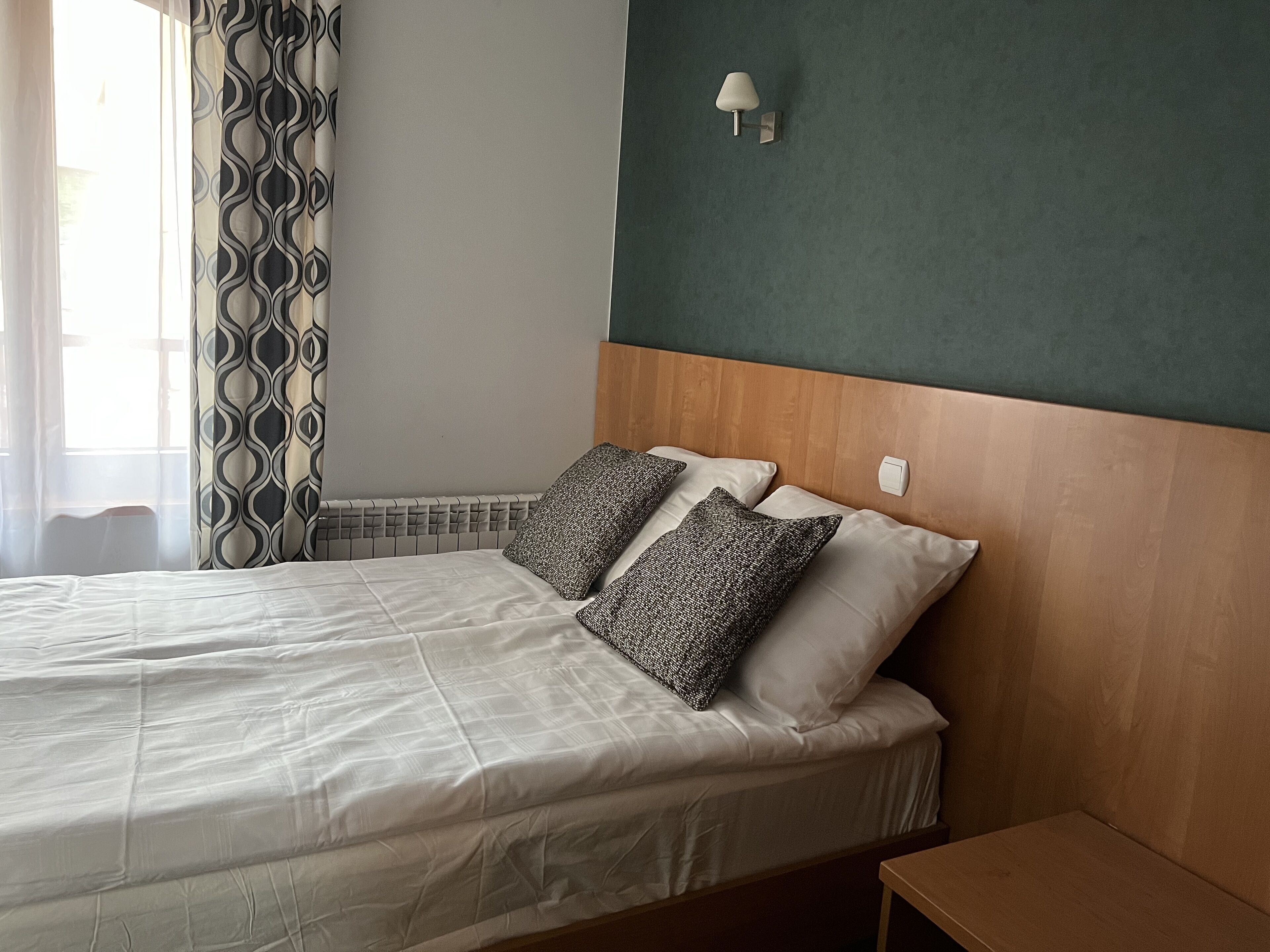 A neatly made bed with white linens and patterned pillows, a wooden headboard, and a wall-mounted light fixture.