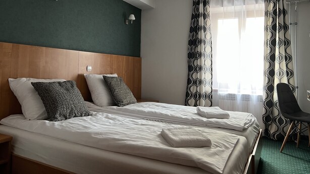 A hotel room with two beds, a wooden headboard, a nightstand, a chair, and a window with patterned curtains. - Garbarska 22 Aparthotel (Kraków)