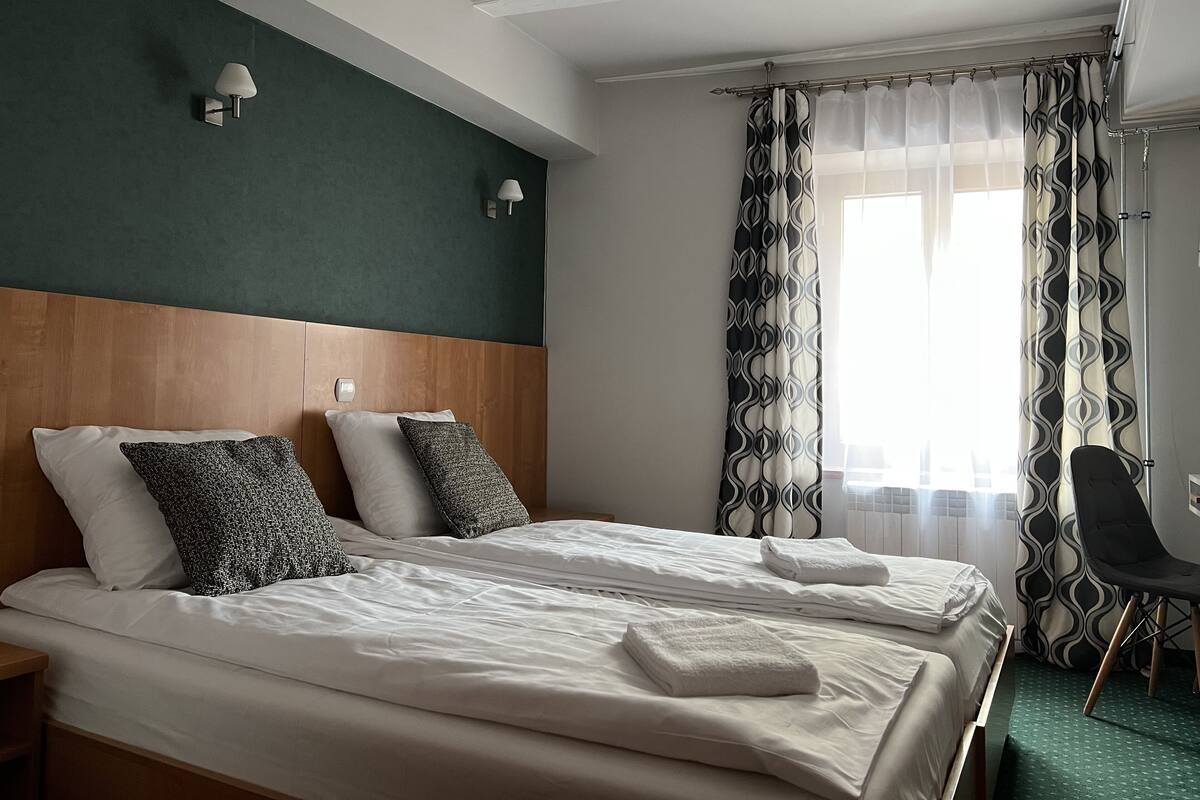 A hotel room with two beds, a wooden headboard, a nightstand, a chair, and a window with patterned curtains.