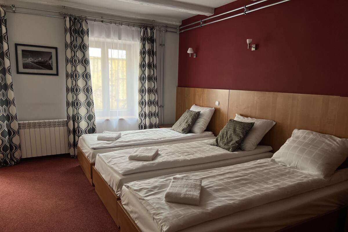 A hotel room with two single beds, a wooden headboard, a red accent wall, a framed picture on the wall, and a window with patterned curtains.