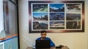 A person sitting at a wooden table with a laptop, in front of a wall with a framed collage of travel photos. - Hotel Royal Mantra (Kathmandu)