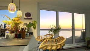 A modern living room with large windows, wicker chairs, and potted plants. - Stunning Sea View Two Bed Apartment - With Balcony (Southbourne)