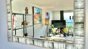 A modern living room with a large mirror reflecting a scenic view, a flat-screen TV, and various decorative items including a sculpture and plants. - Stunning Sea View Two Bed Apartment - With Balcony (Southbourne)