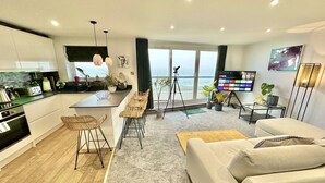A modern kitchen with a central island, a dining area with wicker chairs, a sofa, and a large TV displaying a YouTube interface. - Stunning Sea View Two Bed Apartment - With Balcony (Southbourne)